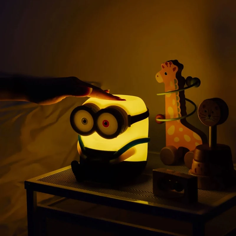 A hand touching the top of the illuminated Minions Bob lamp beside a bed